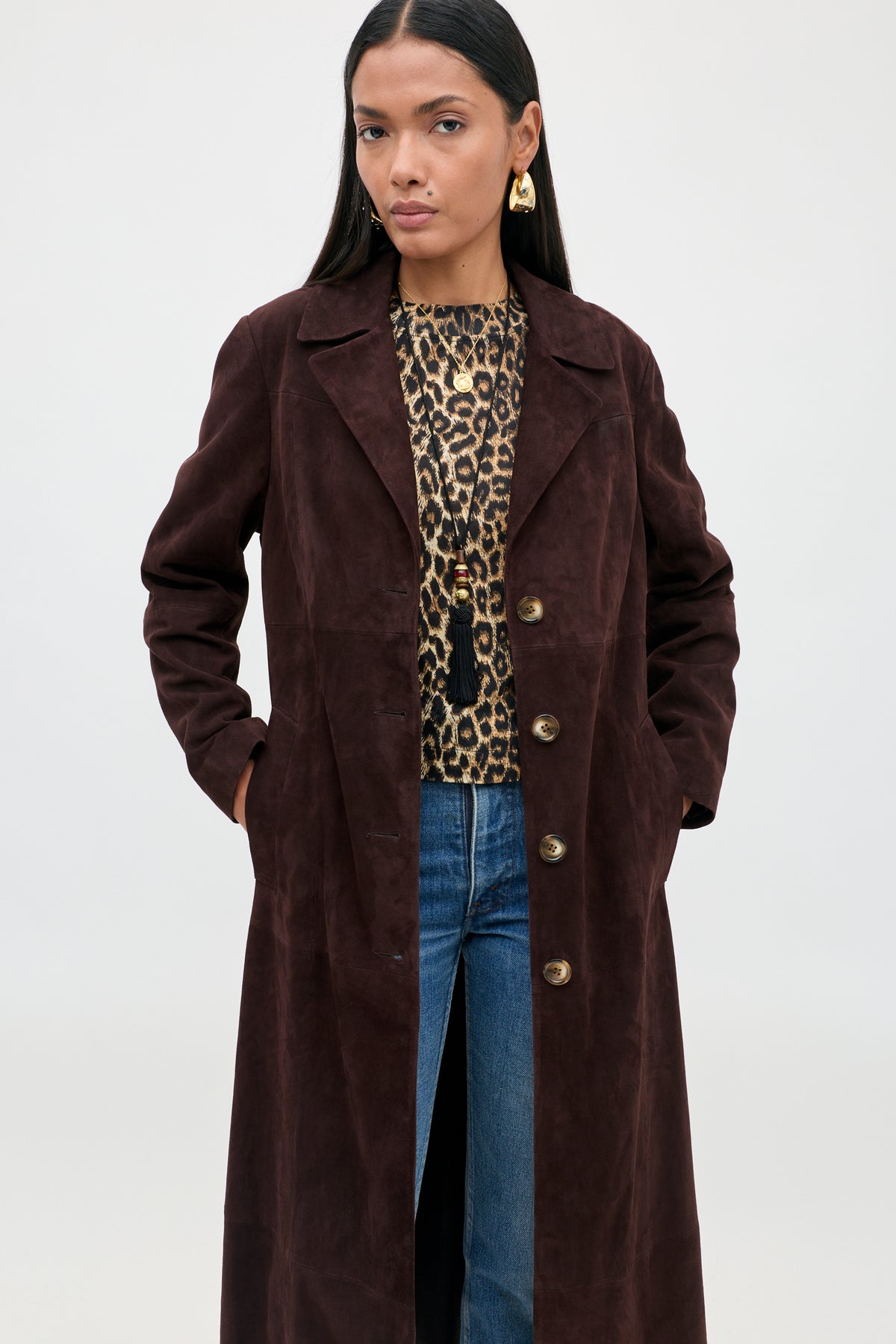 A woman models the Astraea - Chocolate suede coat over a leopard print top and blue jeans, standing with her hands in her coat pockets and looking confidently at the camera against a plain background.