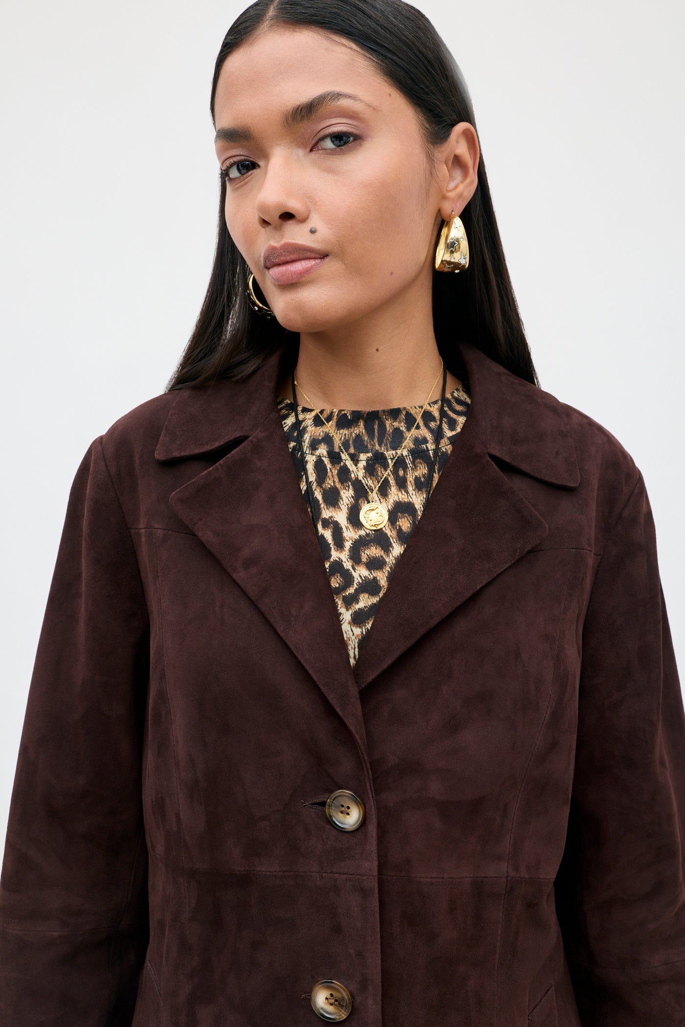 A woman with long dark hair wears gold earrings and the Astraea - Chocolate suede coat with large buttons over a leopard print top. She looks confidently at the camera against a plain light background.