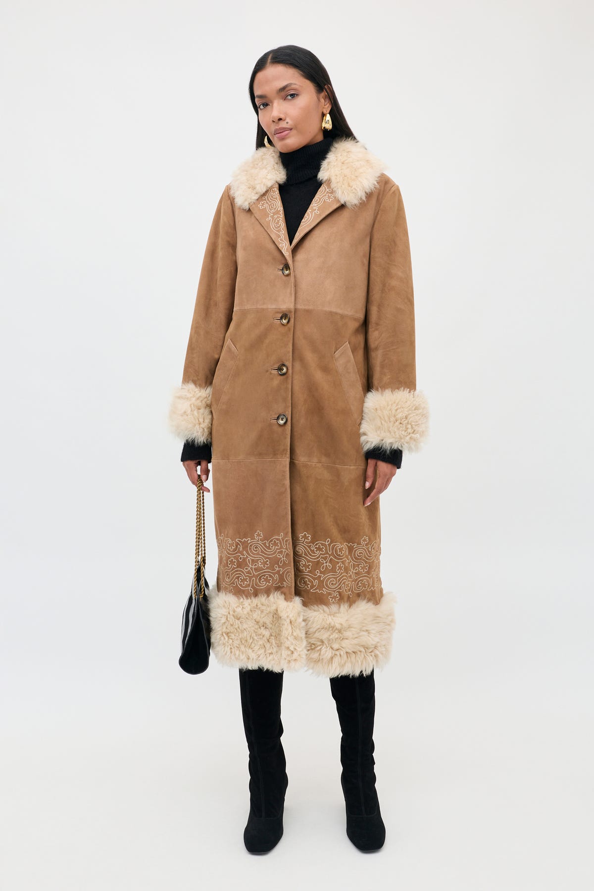 A woman stands against a plain white background wearing the Cienna - Camel long brown suede coat with white fur trim. She pairs the chic and sophisticated coat with a black turtleneck, black pants, black boots, gold earrings, and holds a black handbag. The Cienna - Camel features elegant details that elevate her ensemble with an extra touch of refinement.