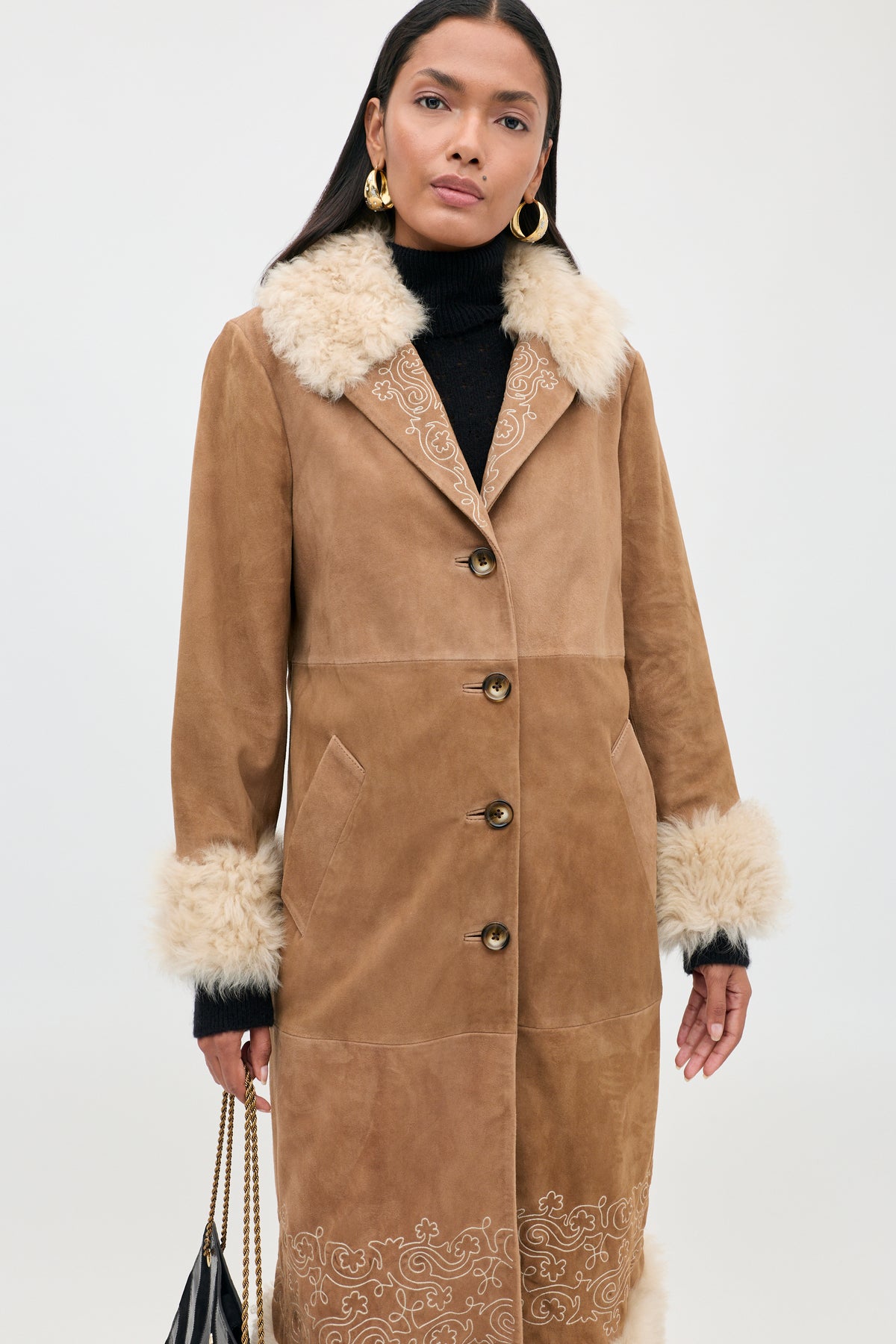 Woman wearing the Cienna - Camel coat, a tan suede piece with a white faux fur collar and cuffs, paired with gold hoop earrings and a black handbag. This chic shearling coat features button closure and decorative embroidery near the hem.