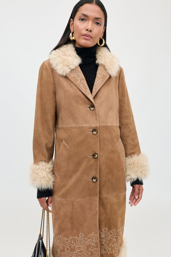 Woman wearing the Cienna - Camel coat, a tan suede piece with a white faux fur collar and cuffs, paired with gold hoop earrings and a black handbag. This chic shearling coat features button closure and decorative embroidery near the hem.