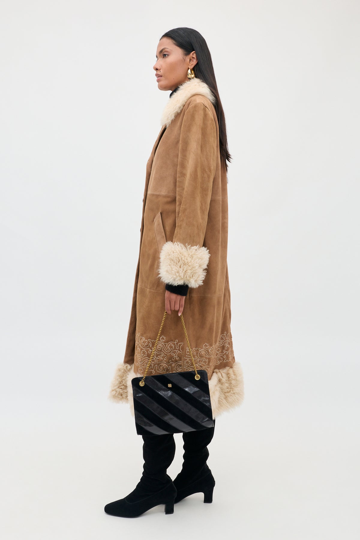 A woman stands sideways against a neutral background, wearing the Cienna - Camel suede coat with fur trim, paired with black leggings and black boots. She carries a black and gold handbag and accessorizes with gold earrings. Her long hair and the elegant Cienna - Camel coat elevate her chic ensemble.