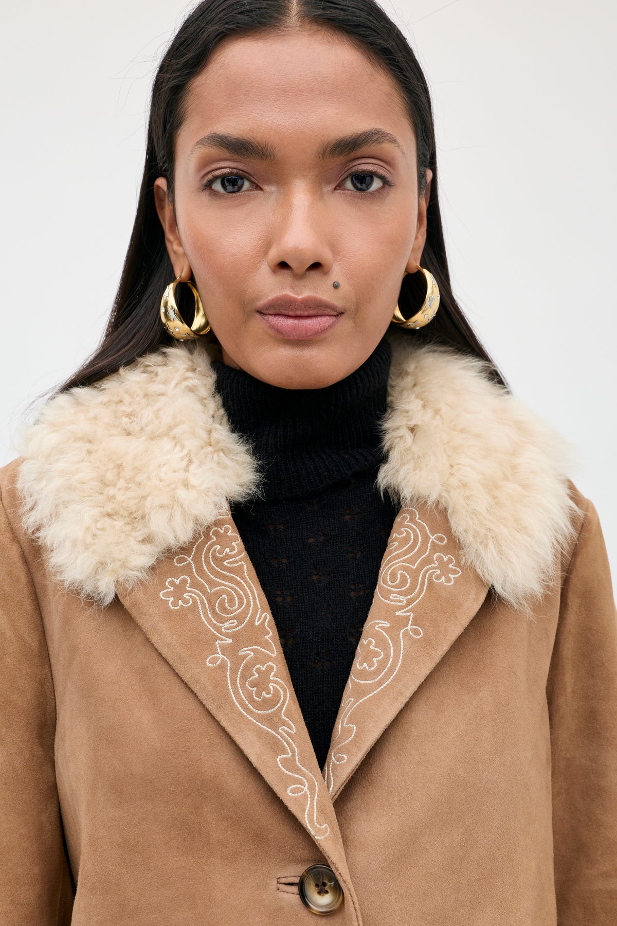 A person wearing the Cienna - Camel shearling coat with a white fur collar, paired with a black turtleneck and large gold hoop earrings, looks directly at the camera against a plain background.