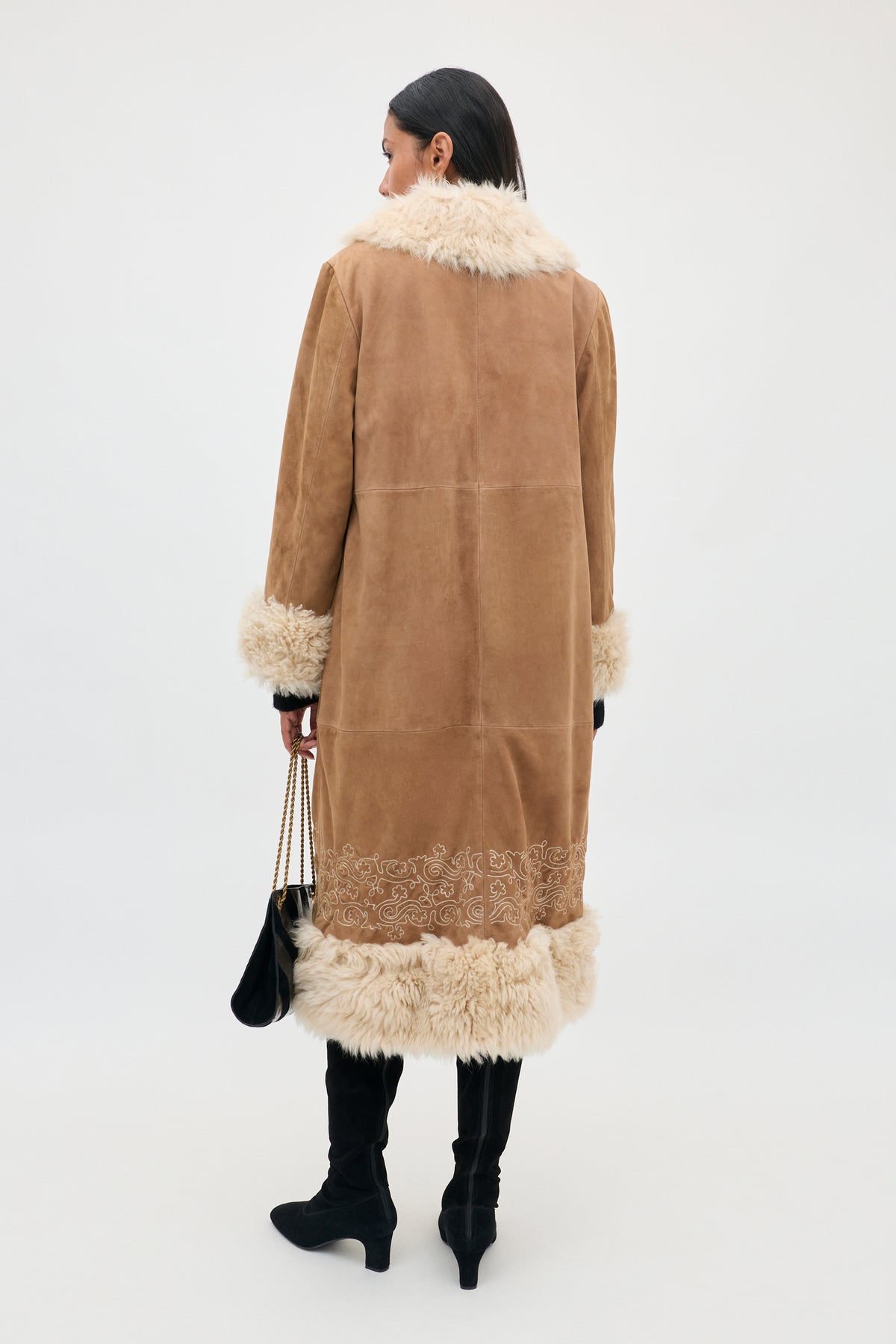 Wearing the Cienna - Camel long brown suede coat with cream-colored fur trim on the collar, cuffs, and hem, a person stands facing away, holding a black handbag and sporting black knee-high boots.