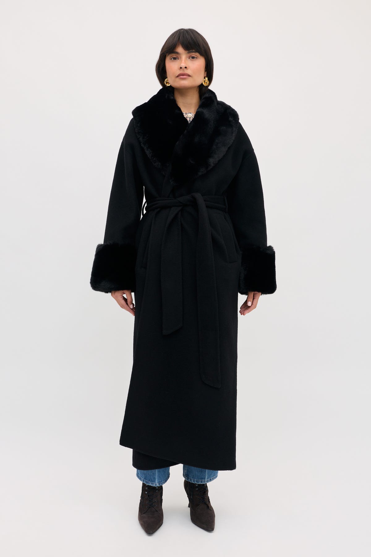 A person wearing a Winslow - Black belted coat with faux fur trim on the collar and cuffs over jeans and heeled boots.