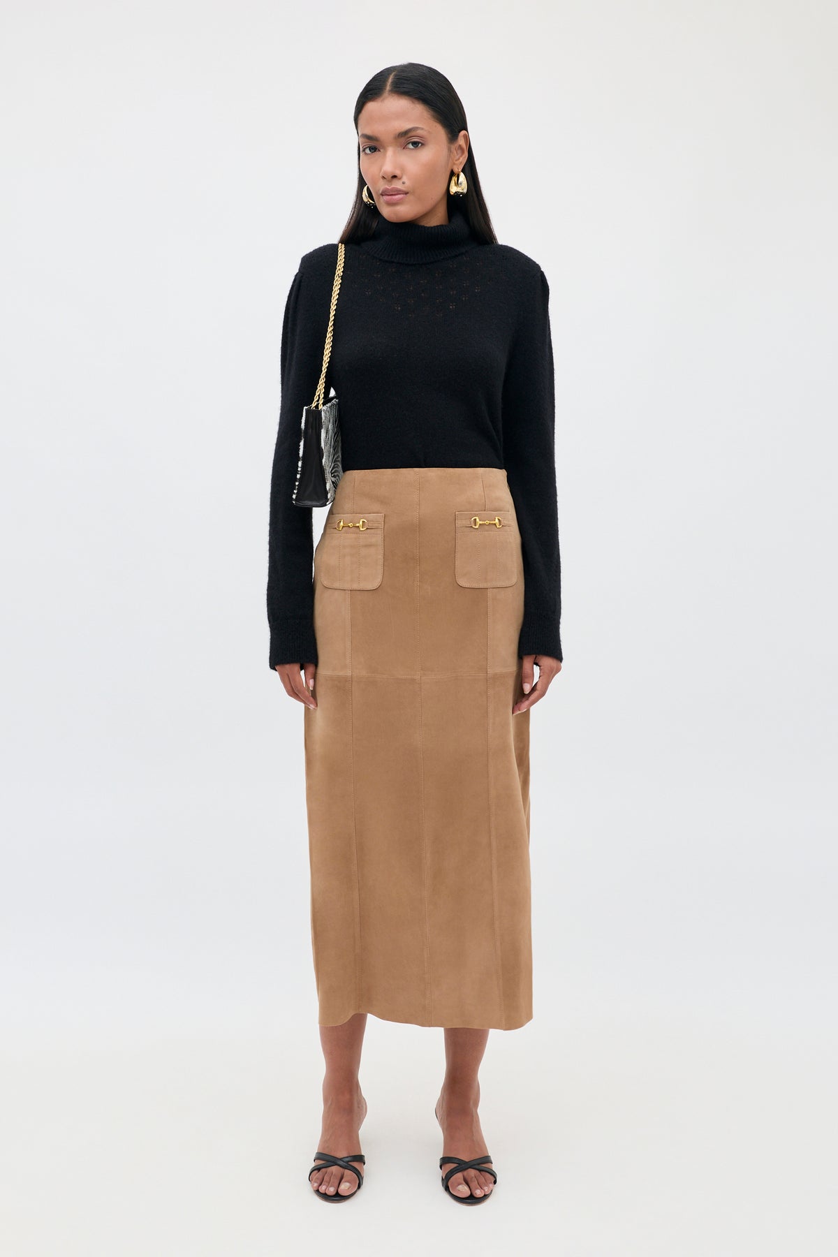 A woman stands facing forward, wearing a black turtleneck sweater, the Edwina - Camel vintage-inspired high-waisted A-line tan suede skirt with front pockets, black sandals, gold earrings, and a black shoulder bag with a gold chain strap.