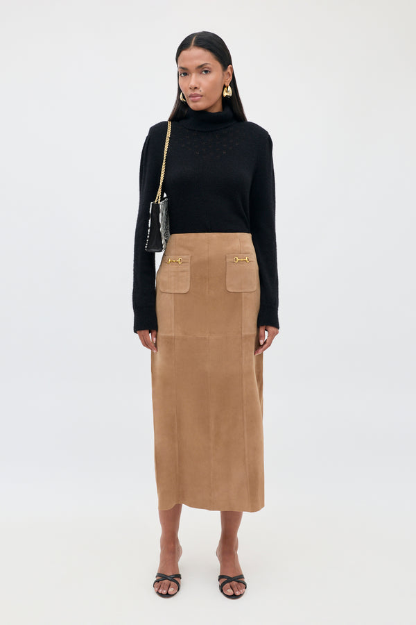 A woman stands facing forward, wearing a black turtleneck sweater, the Edwina - Camel vintage-inspired high-waisted A-line tan suede skirt with front pockets, black sandals, gold earrings, and a black shoulder bag with a gold chain strap.