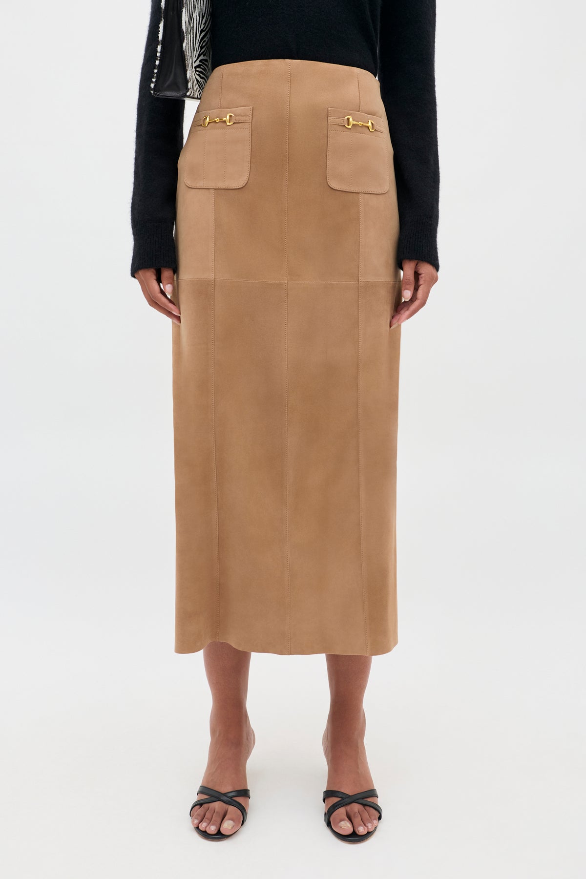 Wearing the Edwina - Camel skirt, which features a vintage-inspired camel suede design in a high-waisted A-line midi cut with front pockets and gold accents, the person pairs it with a black long-sleeve top and black sandals against a plain white background.