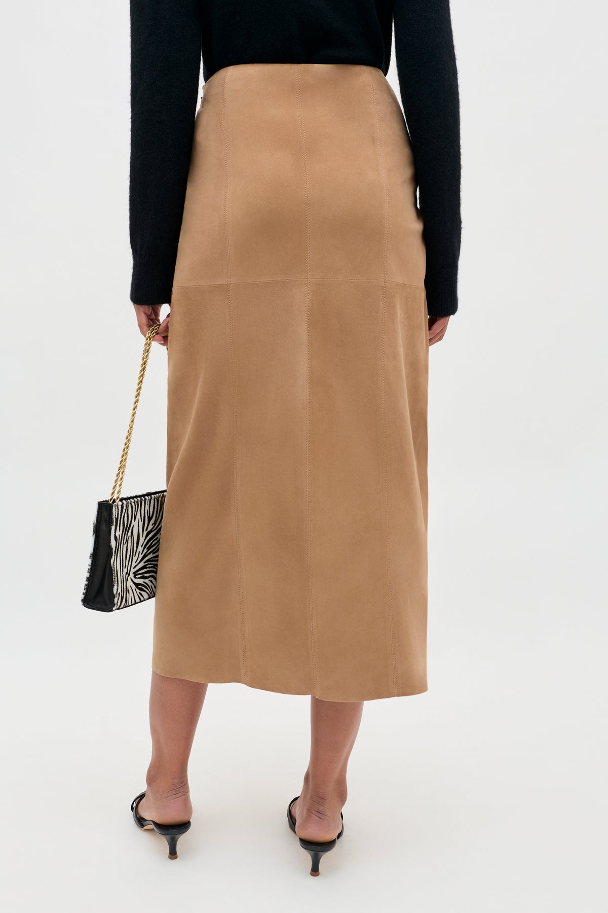 A person is seen from behind against a plain background, wearing the Edwina - Camel high-waisted A-line tan midi skirt, paired with a black long-sleeve top, black heeled sandals, and holding a small zebra print purse with a gold chain.