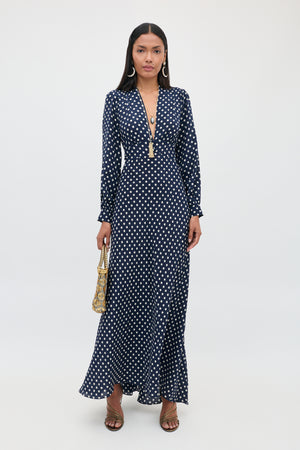 A woman stands against a plain white background wearing the Emory - Runway Spot Navy dress, which features long sleeves, a deep V-neck, and navy blue polka dots; she accessorizes with a gold handbag, long earrings, and sandals.