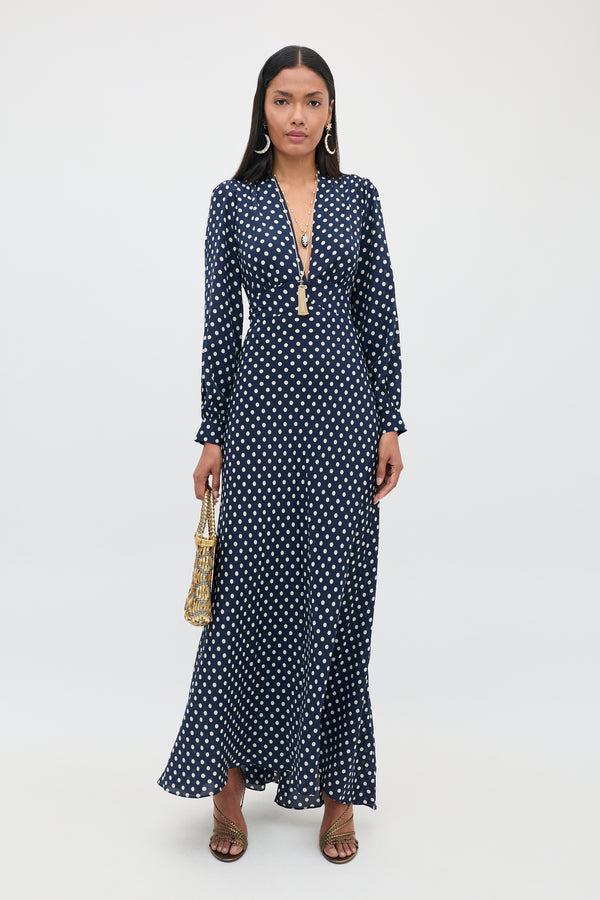 A woman stands against a plain white background wearing the Emory - Runway Spot Navy dress, which features long sleeves, a deep V-neck, and navy blue polka dots; she accessorizes with a gold handbag, long earrings, and sandals.