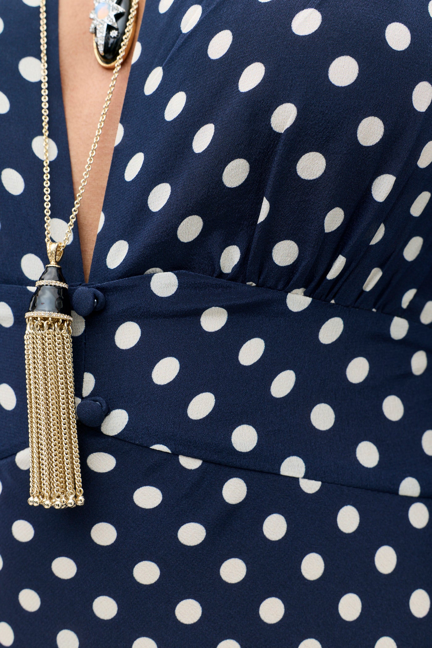 Close-up of the Emory - Runway Spot Navy dress, showcasing its signature deep V-neckline and classic navy polka dot pattern. The outfit is tastefully accessorized with a gold tassel necklace and an additional necklace featuring a dark star-adorned pendant.