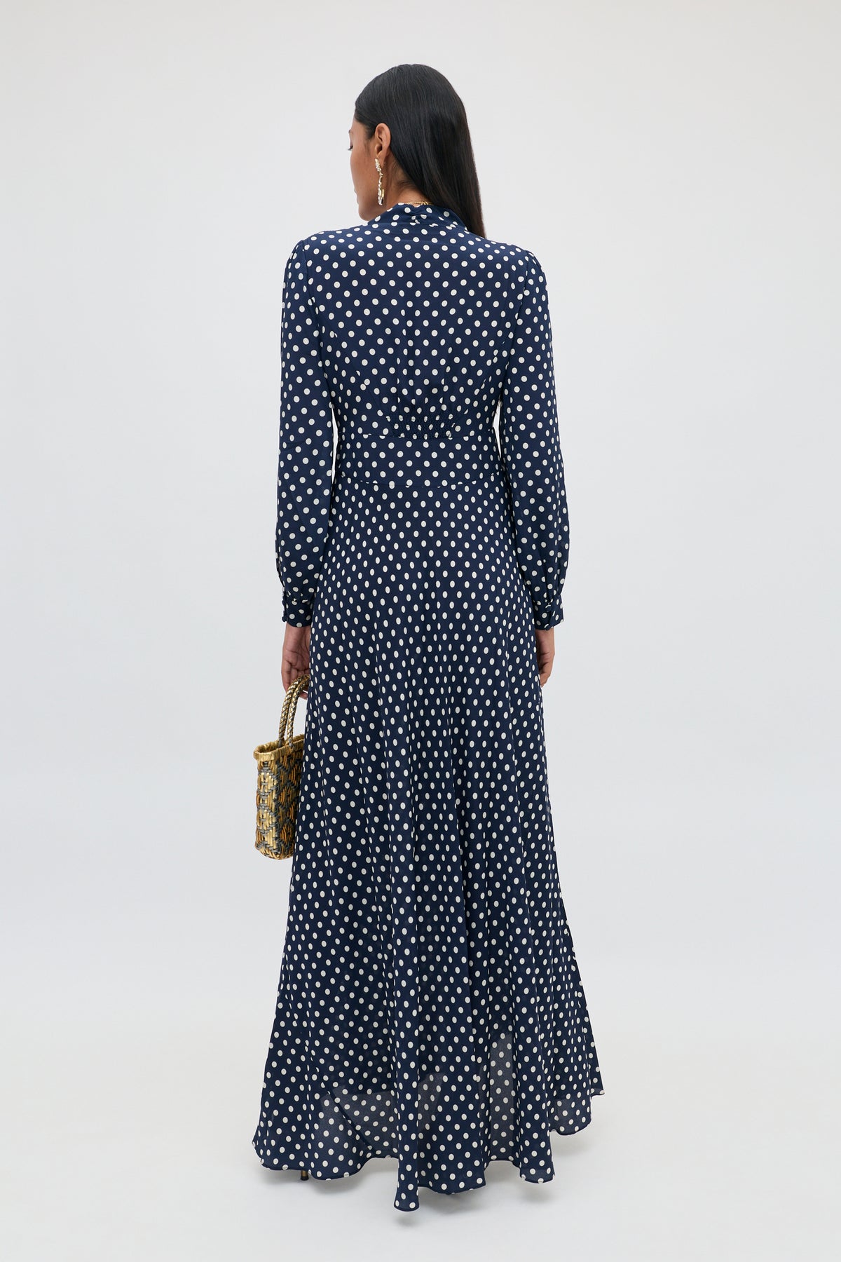 A person with long dark hair, wearing the Emory - Runway Spot Navy dress—a navy blue, floor-length, long-sleeve dress with polka dots—is standing and facing away while holding a small woven handbag.
