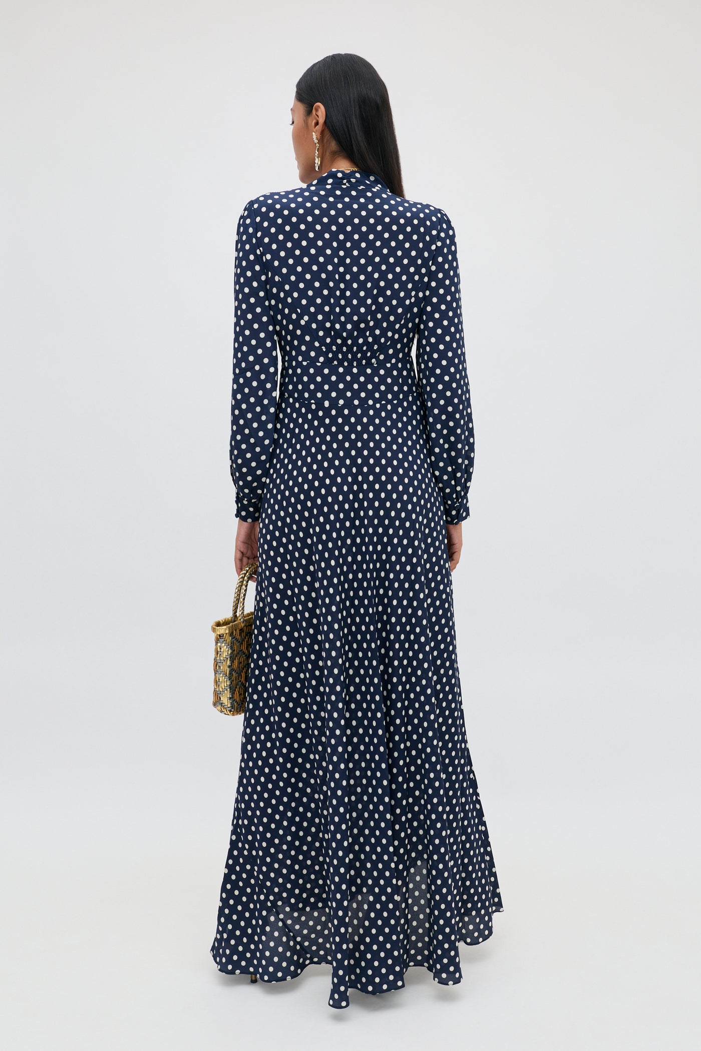 A person with long dark hair, wearing the Emory - Runway Spot Navy dress—a navy blue, floor-length, long-sleeve dress with polka dots—is standing and facing away while holding a small woven handbag.