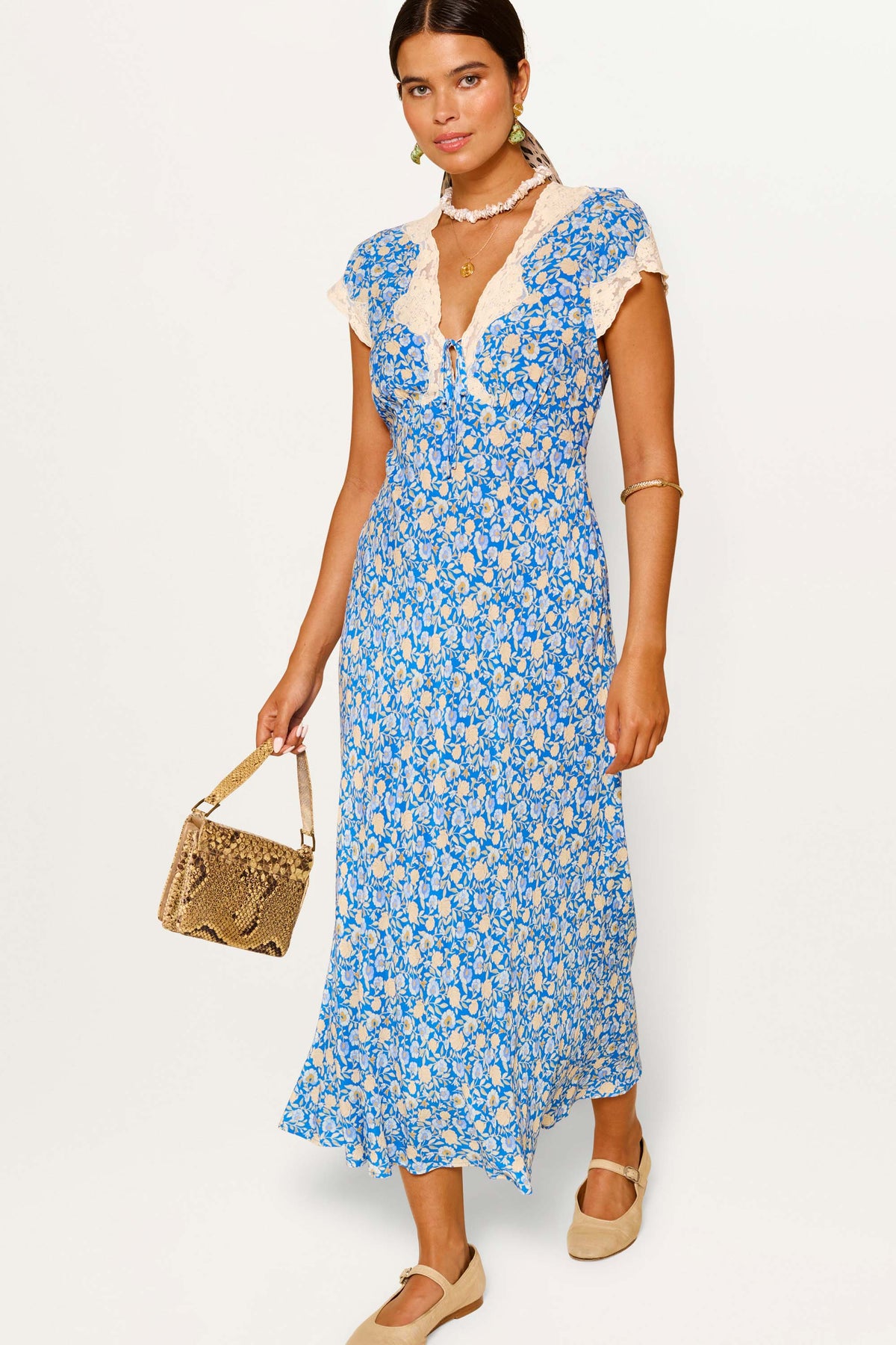 A woman in the Amari dress, Shell Pansy Indigo print, with a bias silhouette, holds a brown woven handbag and wears gold jewelry and beige shoes, standing against a plain white background.