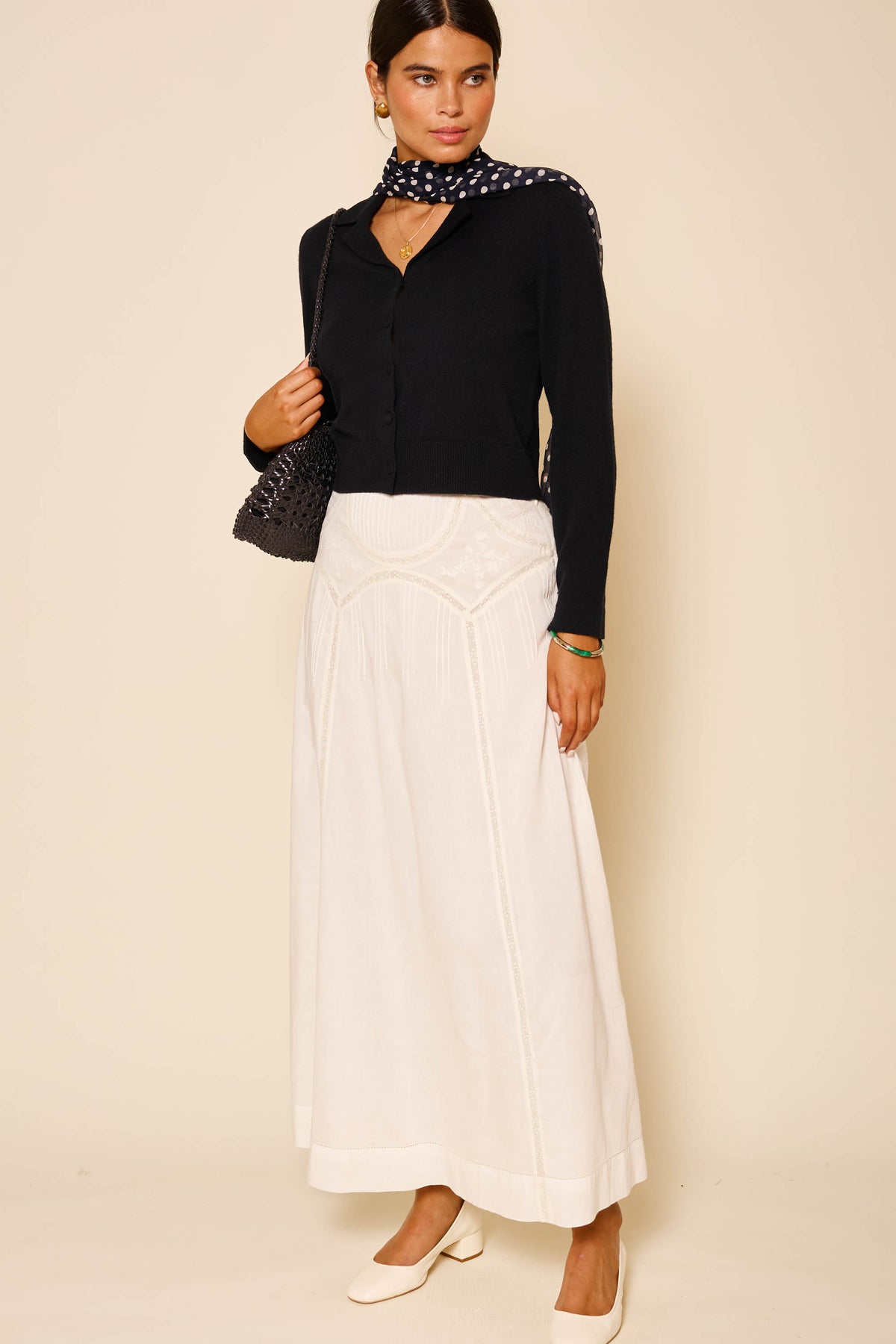A woman stands against a beige background wearing a black RIXO Carla cardigan, white maxi skirt, white shoes, a black polka dot scarf, and carries a black bag over her shoulder.
