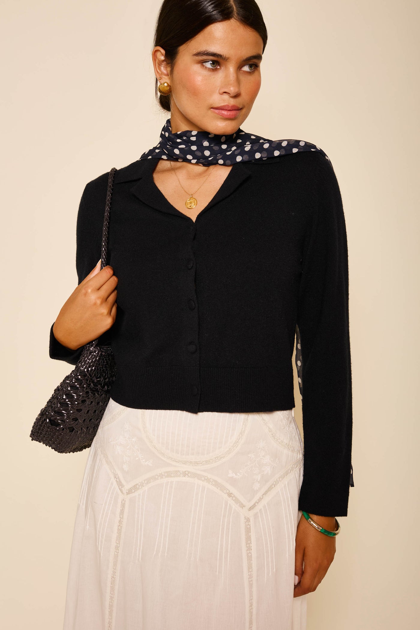 A woman stands against a plain background wearing Carla - Black, a RIXO wool cashmere blend cardigan, with a white skirt, black shoulder bag, polka dot scarf, gold jewelry, and her dark hair pulled back.
