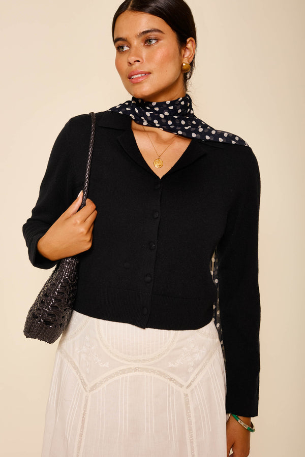 A woman wears a black wool cashmere blend cardigan, a white embroidered skirt, a black polka dot scarf, gold earrings, a pendant necklace, and carries RIXO’s textured black “Carla - Black” shoulder bag.