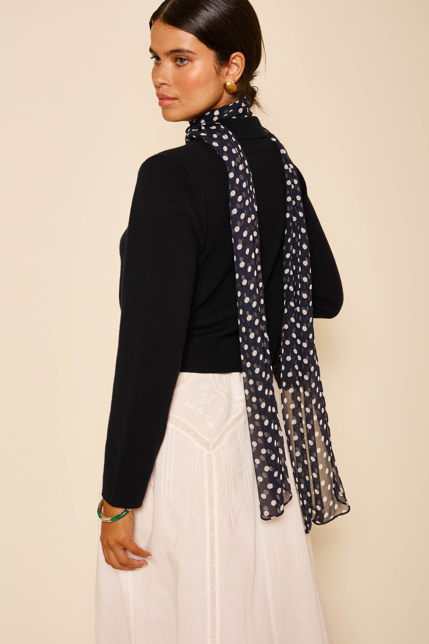 A woman stands sideways, wearing the Carla - Black wool cashmere turtleneck sweater, paired with a long white embroidered skirt and a sheer black scarf with white polka dots. Her dark hair is pulled back and she accessorizes with gold earrings.