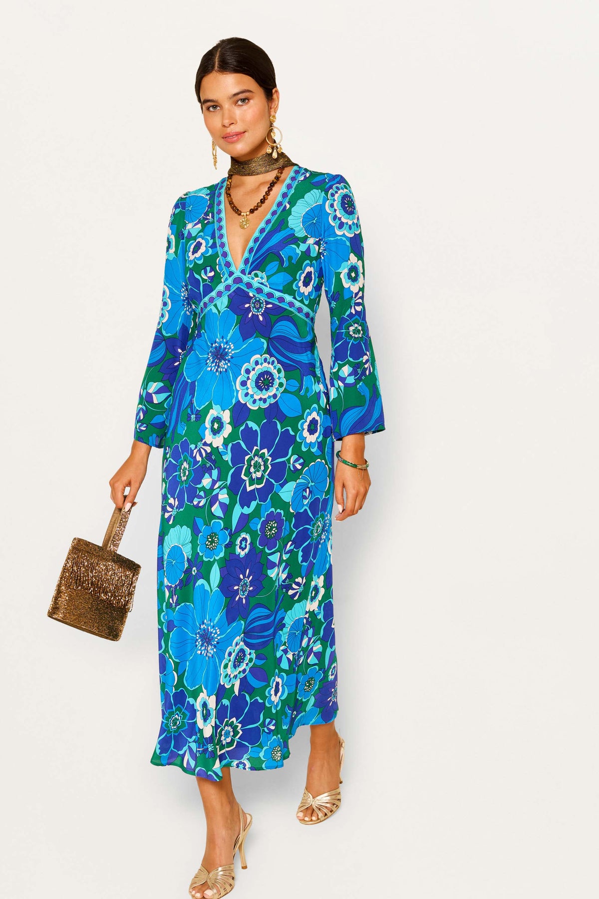 Woman wearing the tania - miami floral emerald, a long, green silk gown with a V-neck and long sleeves, styled with layered necklaces, gold earrings, gold heels, and a gold textured handbag.