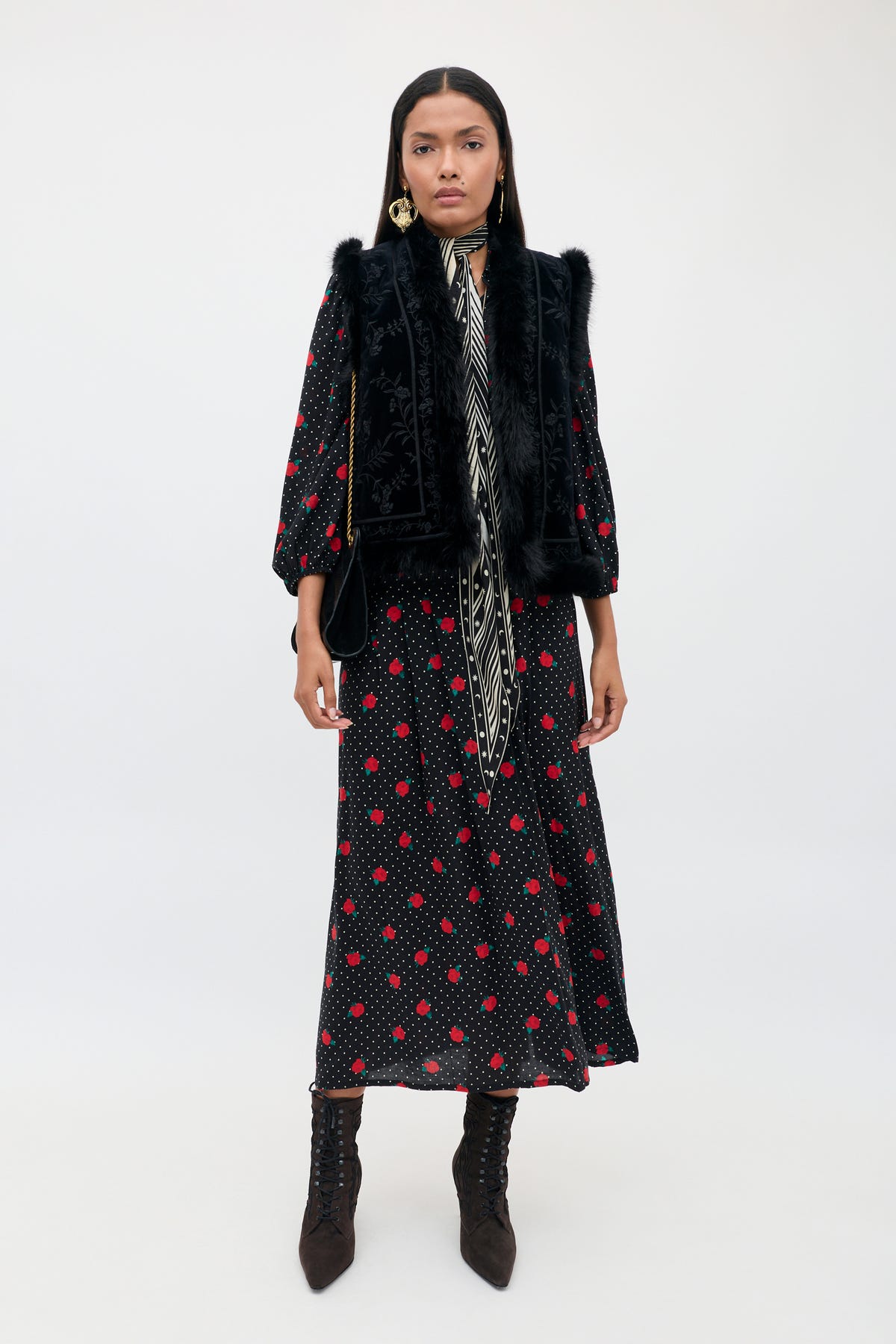A woman faces forward against a plain white background, wearing the Izzy - Rose Spot Black dress—a long black dress featuring red polka dots and patterned stripes—styled with a black fur vest, black heeled boots, gold earrings, and her long dark hair worn loose.