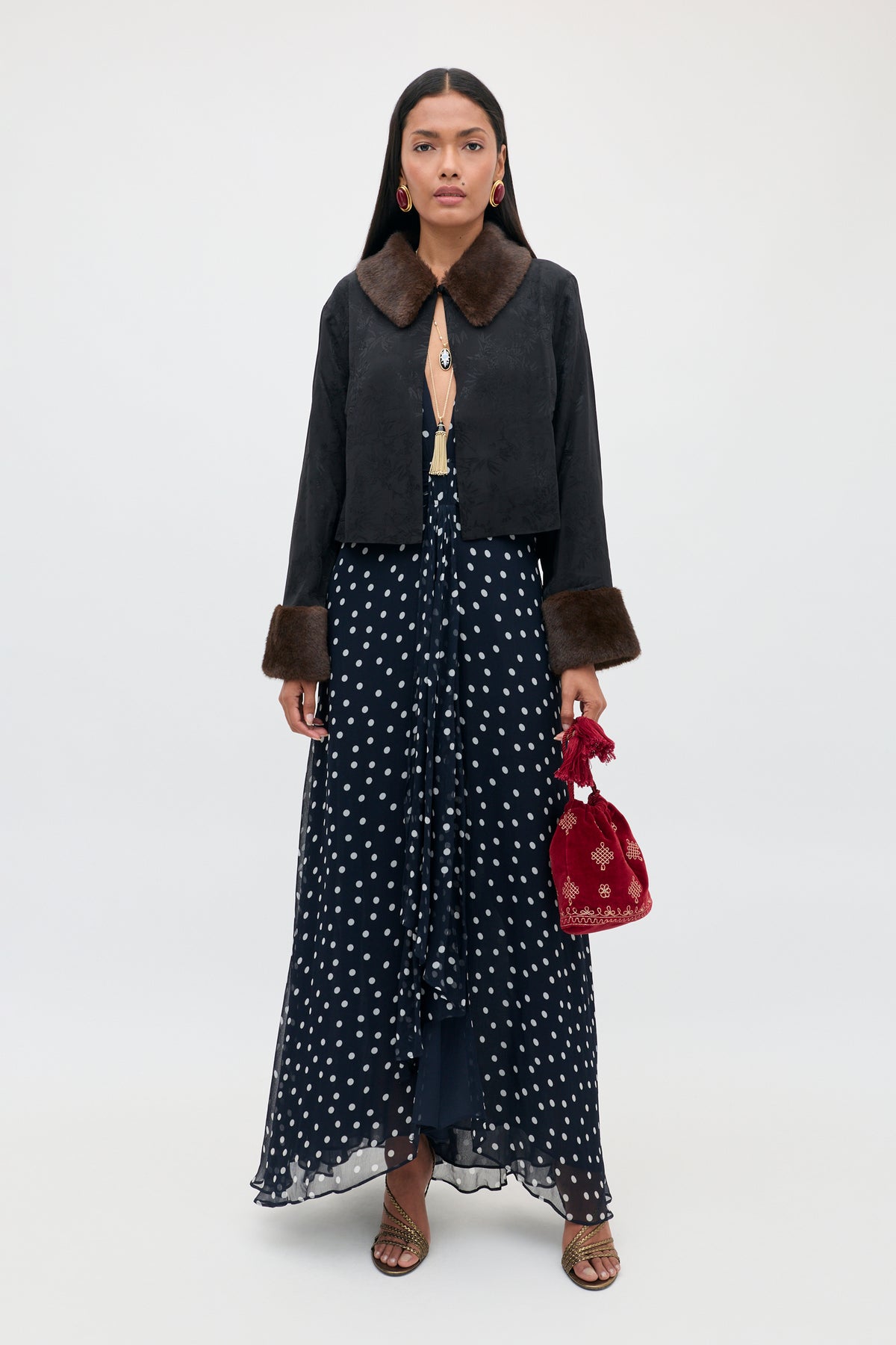 A woman stands facing forward wearing the Meera - Bohemia Spot Deep Navy gown, styled with a black faux fur-trimmed jacket, gold sandals, red earrings, and a small red embroidered bag against a plain background.