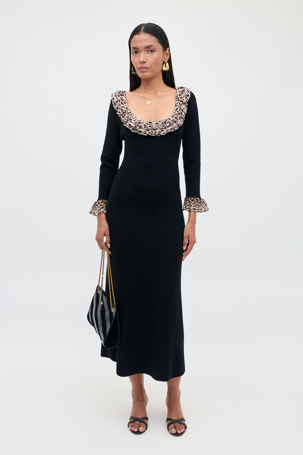 A woman wearing the Mika - Antelope Spot Black, a long black merino wool knit dress with ruffled leopard print trim at the neckline and cuffs, stands against a plain white background. She pairs the dress with black sandals, hoop earrings, and carries a black handbag.