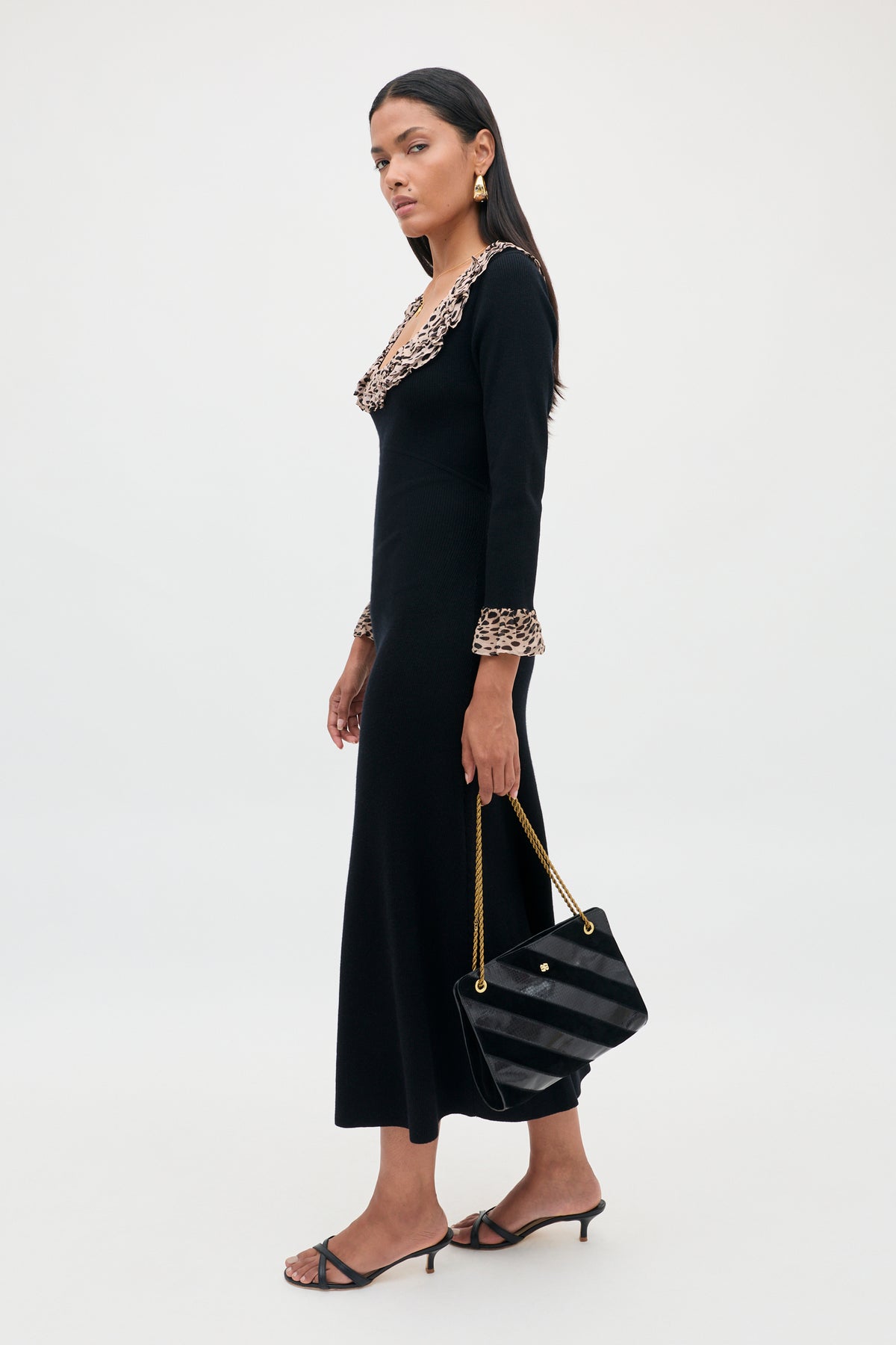 A woman wearing the Mika - Antelope Spot Black long dress with a scoop neckline and ruffled leopard trim, styled with black heeled sandals, gold earrings, and a black chain-strap handbag, stands against a plain white background.