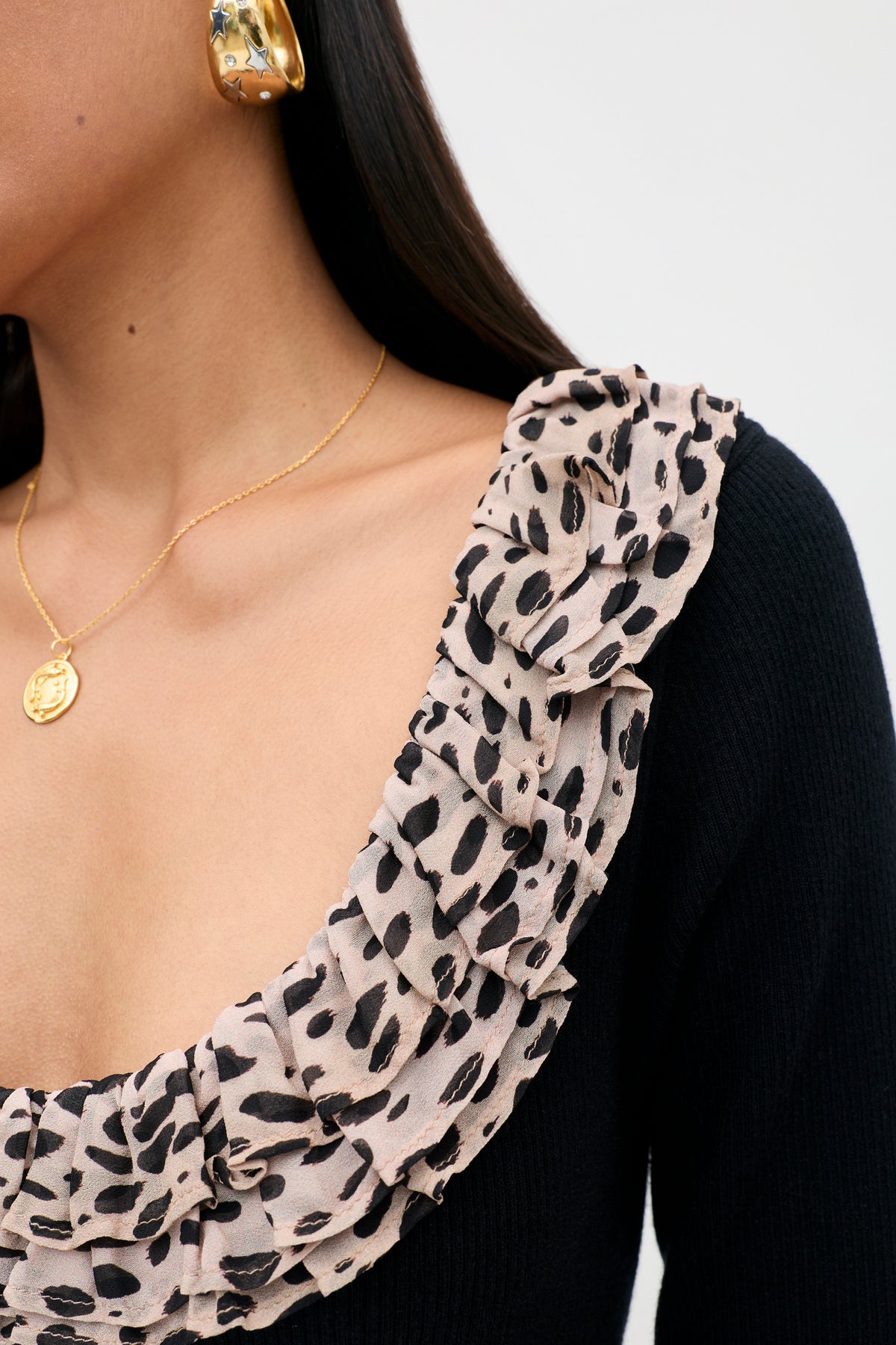 Close-up of a person wearing a gold earring and gold necklace, dressed in the Mika - Antelope Spot Black top featuring a beige, ruffled leopard trim neckline. The background is plain and light-colored.