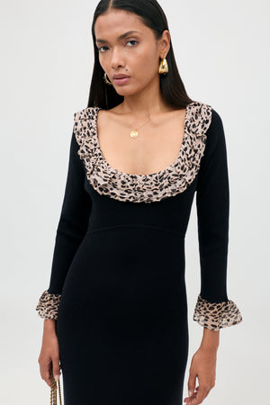 A woman wearing the Mika - Antelope Spot Black dress, featuring a black scoop neckline with ruffled leopard-spot trim at the neckline and cuffs, styled with gold earrings and a gold necklace, stands against a plain light background.