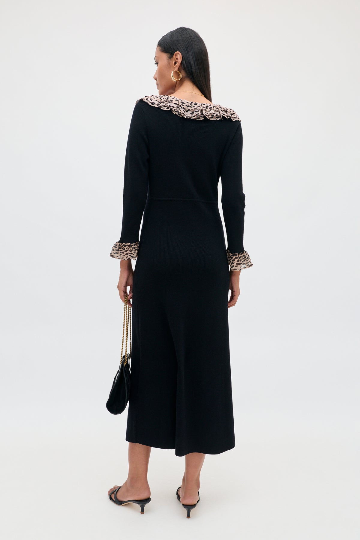 A woman stands facing away, wearing the Mika - Antelope Spot Black dress—a long black scoop neck style featuring ruffled leopard trim at the collar and cuffs. She has long dark hair, wears hoop earrings, holds a black purse, and pairs the look with black heeled sandals against a light background.