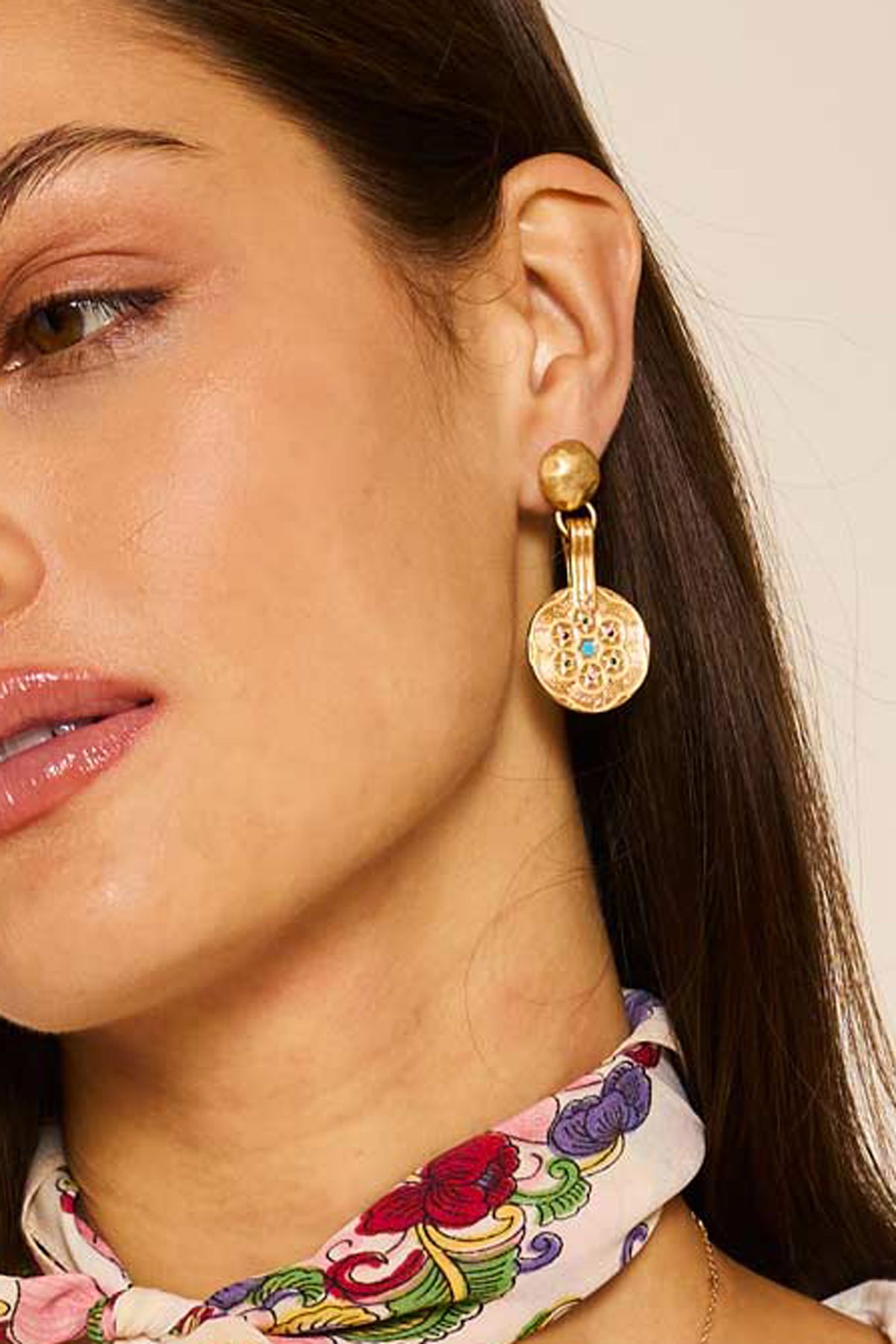 A woman with brown hair tucked behind her ear wears a colorful floral scarf and the Anouk - Gold earrings, featuring circular, textured designs and small gemstones; shown from the lips up.