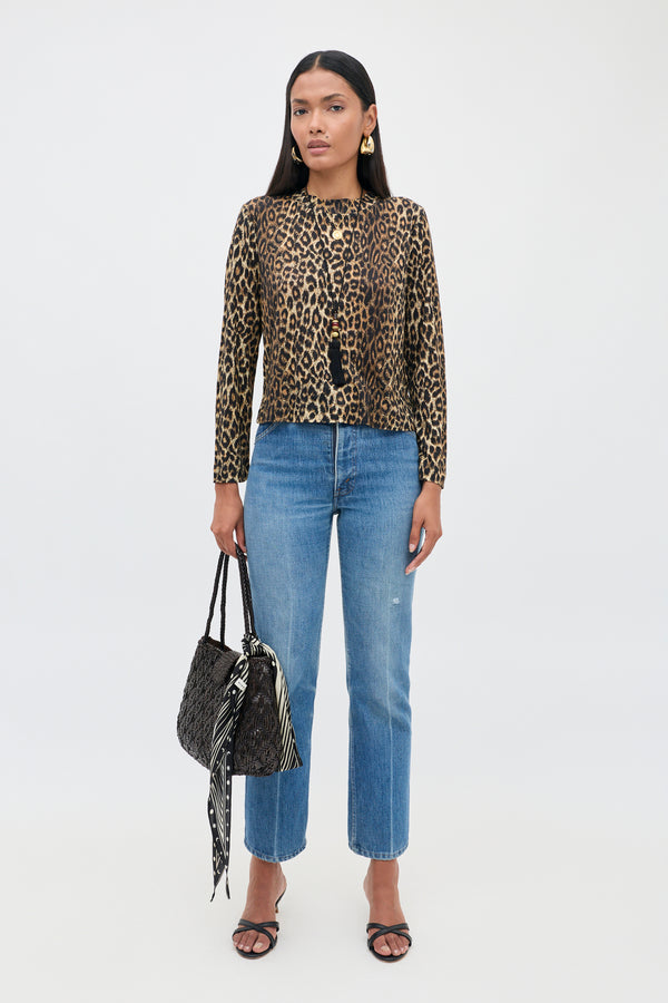 A woman stands against a plain background, wearing the Phaedra - Lyra Leopard Black long-sleeve top crafted from soft Tencel Jersey, paired with blue jeans and black heeled sandals. She holds a black purse adorned with a tied scarf.