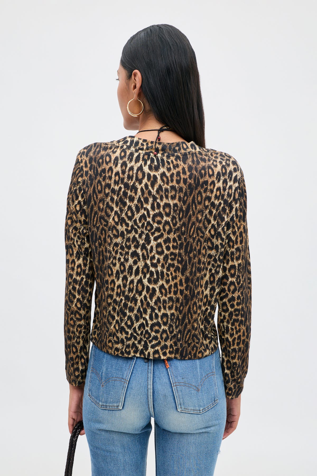 A woman with long dark hair, wearing hoop earrings and the Phaedra - Lyra Leopard Black long-sleeve crop top made from soft Tencel Jersey, paired with blue jeans, stands with her back to the camera against a plain light background.