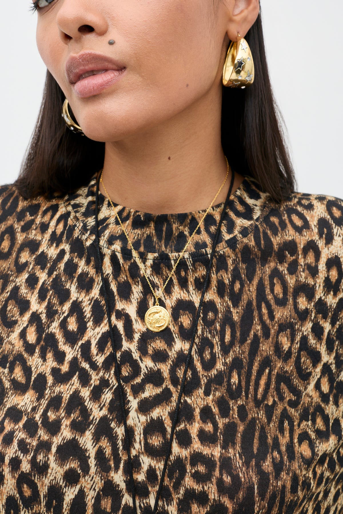A person wearing the Phaedra - Lyra Leopard Black crop top, crafted from soft Tencel Jersey, paired with large gold earrings and layered gold necklaces. Only the lower face and upper torso are visible.