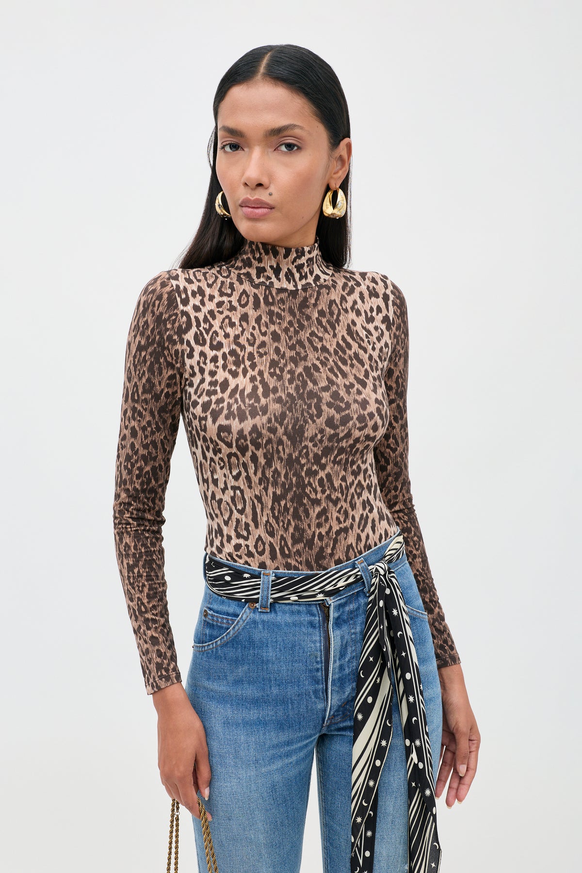 A woman stands against a plain light background wearing the Polo - Leopard Brown bodysuit crafted from soft TENCEL™ Modal, paired with blue jeans, gold hoop earrings, and a patterned scarf tied at her waist.
