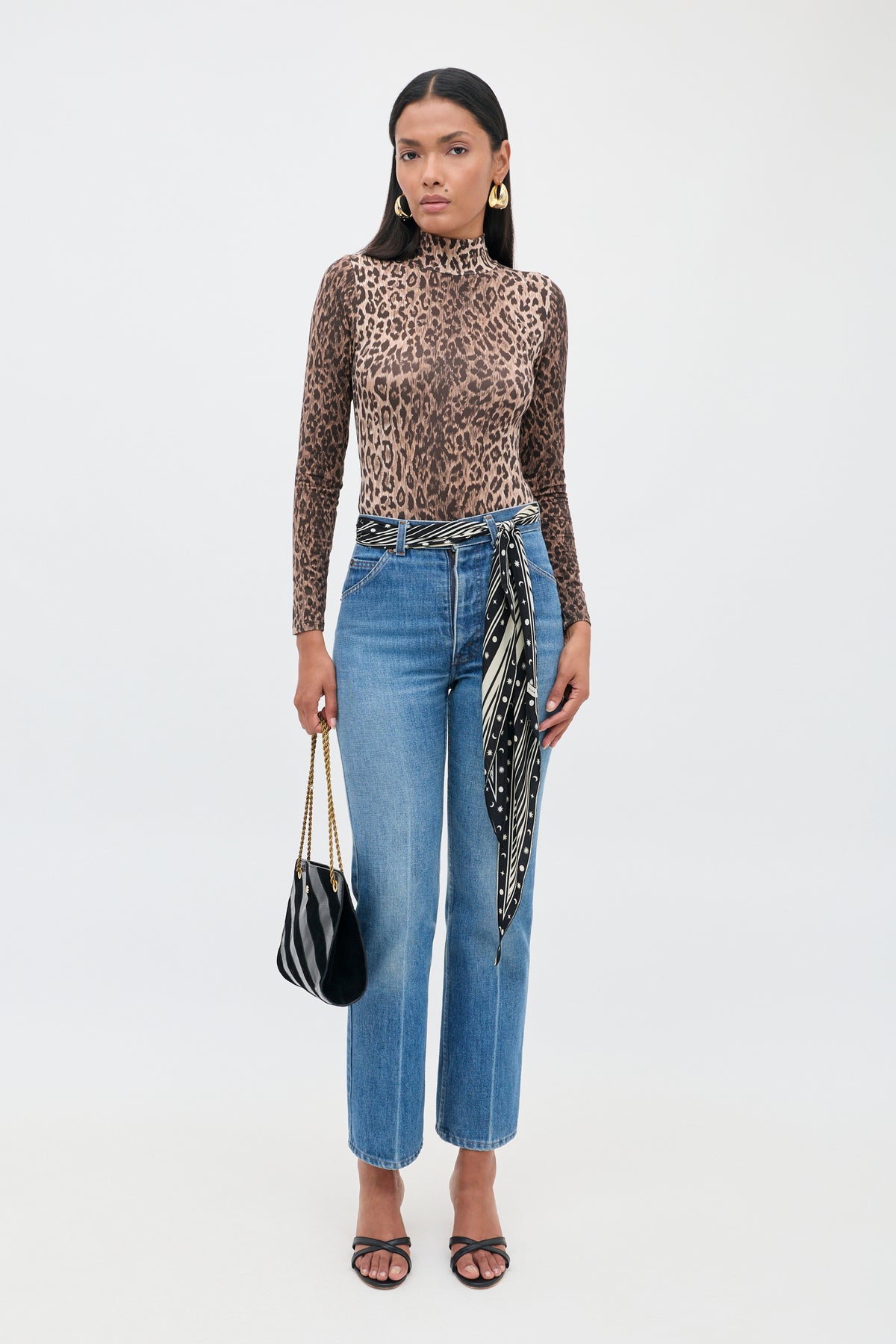 A woman stands facing forward against a plain white background, wearing the Polo - Leopard Brown bodysuit, blue jeans styled with a black-and-white scarf belt, black sandals, gold earrings, and holding a black purse with a chain strap.