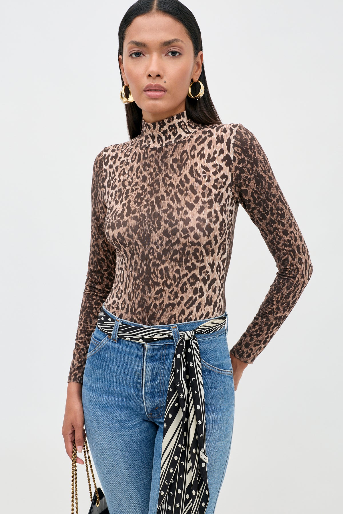 Woman wearing the Polo - Leopard Brown, a turtleneck top crafted from soft TENCEL™ Modal, styled with blue jeans featuring a striped scarf belt, gold hoop earrings, and holding a black handbag, standing against a plain white background.