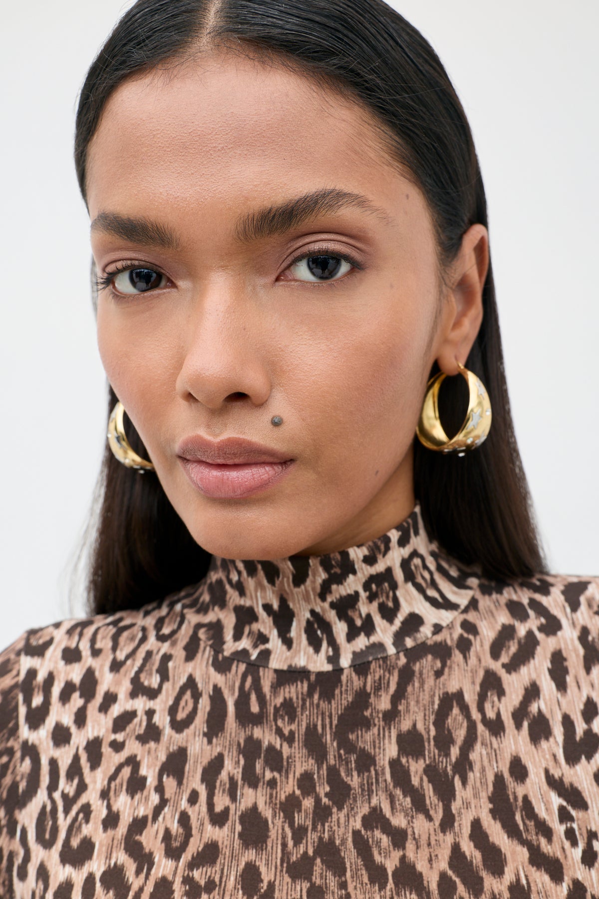 A woman with straight dark hair wears gold hoop earrings and a Polo - Leopard Brown top made from TENCEL Modal, looking directly at the camera against a plain, light-colored background.