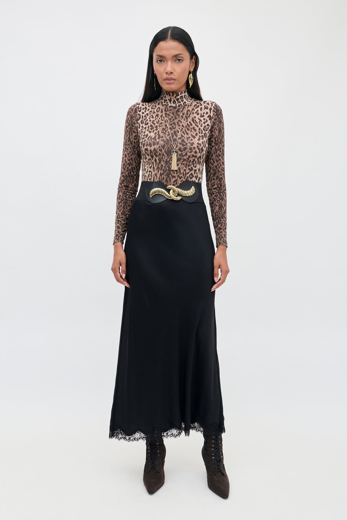 A person wearing the Polo - Leopard Brown long-sleeve top paired with a black high-waisted skirt, snake-shaped belt, gold earrings, and dark boots stands against a plain white background. The striking leopard pattern of the Polo - Leopard Brown lends bold flair to the outfit, while statement accessories highlight the look’s modern silhouette.
