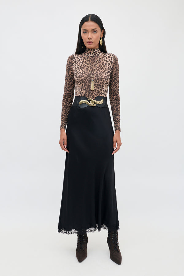 A person wearing the Polo - Leopard Brown long-sleeve top paired with a black high-waisted skirt, snake-shaped belt, gold earrings, and dark boots stands against a plain white background. The striking leopard pattern of the Polo - Leopard Brown lends bold flair to the outfit, while statement accessories highlight the look’s modern silhouette.