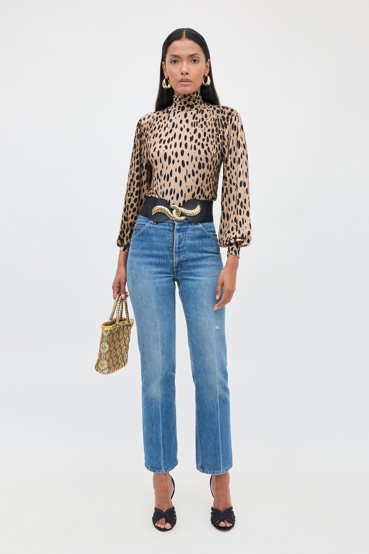 A woman wearing the Suki - Antelope Spot Black top, high-waisted blue jeans, black heeled sandals, a black belt with a gold snake buckle, gold hoop earrings, and carrying a patterned handbag.