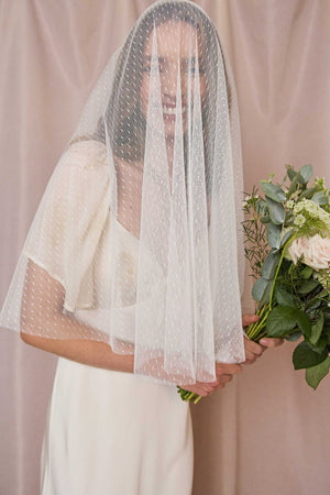 A bride in a white dress smiles, holding a bouquet, with the Cupid - Ivory Veil—an ivory polka-dotted mesh bridal veil—gently draped over her face against a soft beige curtain backdrop.