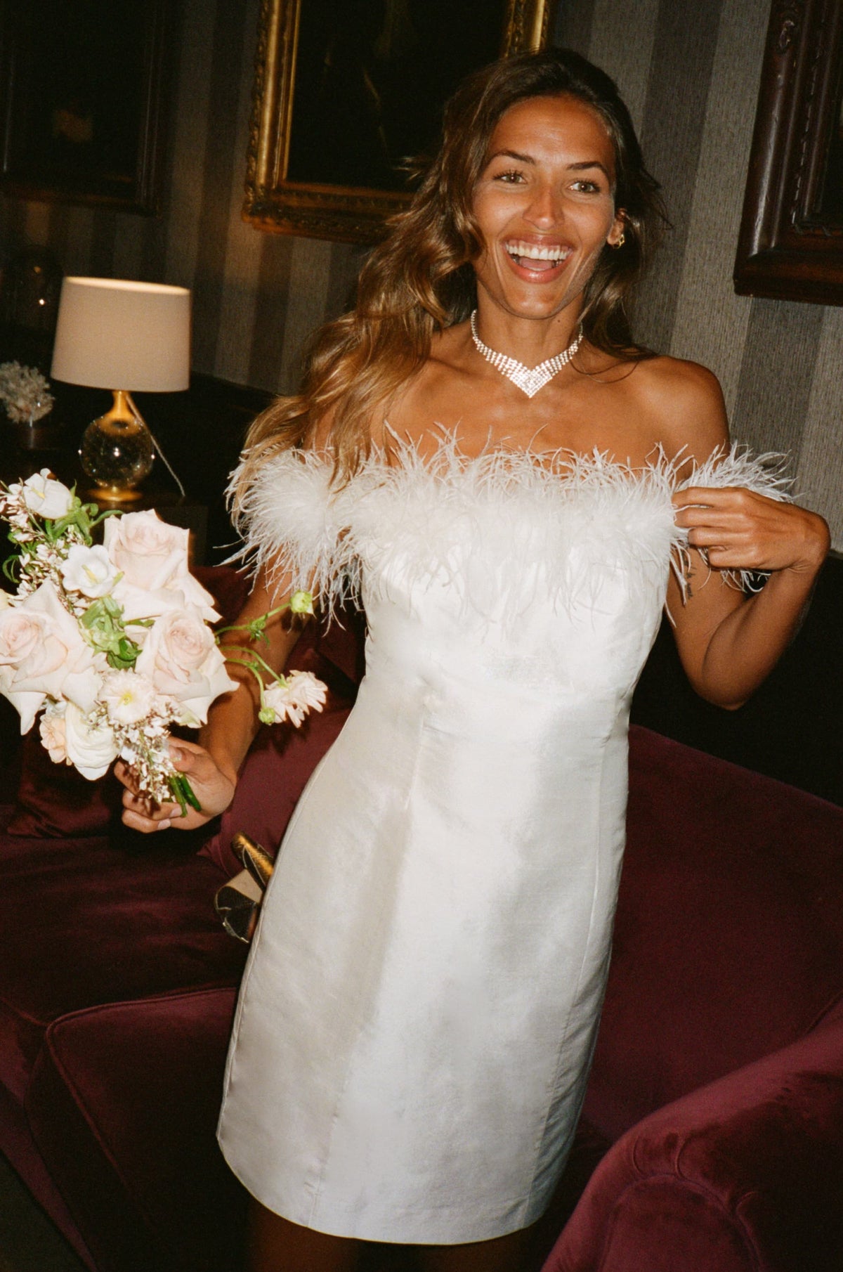 A smiling woman wears the Lena - Feather Ivory dress with Bardot neckline, feather trim, and satin finish, holding light pink roses indoors near a lamp and framed art. She also wears a necklace.