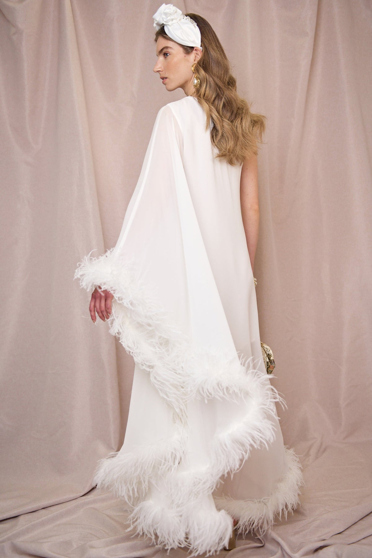 A woman in the Liza - Ivory Feathers silk midi dress with feathered trim stands against a soft beige backdrop, her wavy hair styled with a white headband, gold earrings, and a small clutch as she glances elegantly over her shoulder.