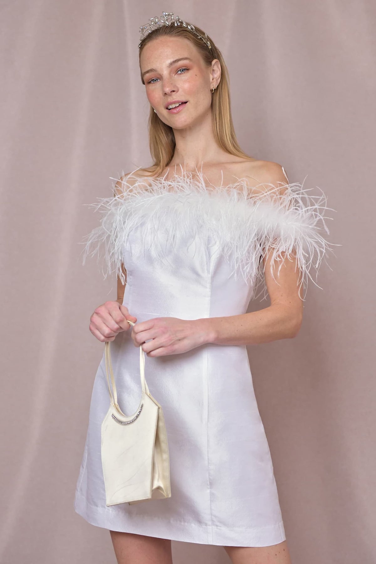 A woman smiles softly at the camera, wearing Lena - Feather Ivory, a white silk-jacquard mini dress with a Bardot neckline and feather trim. She pairs it with a small cream handbag and a silver tiara against a light beige background.