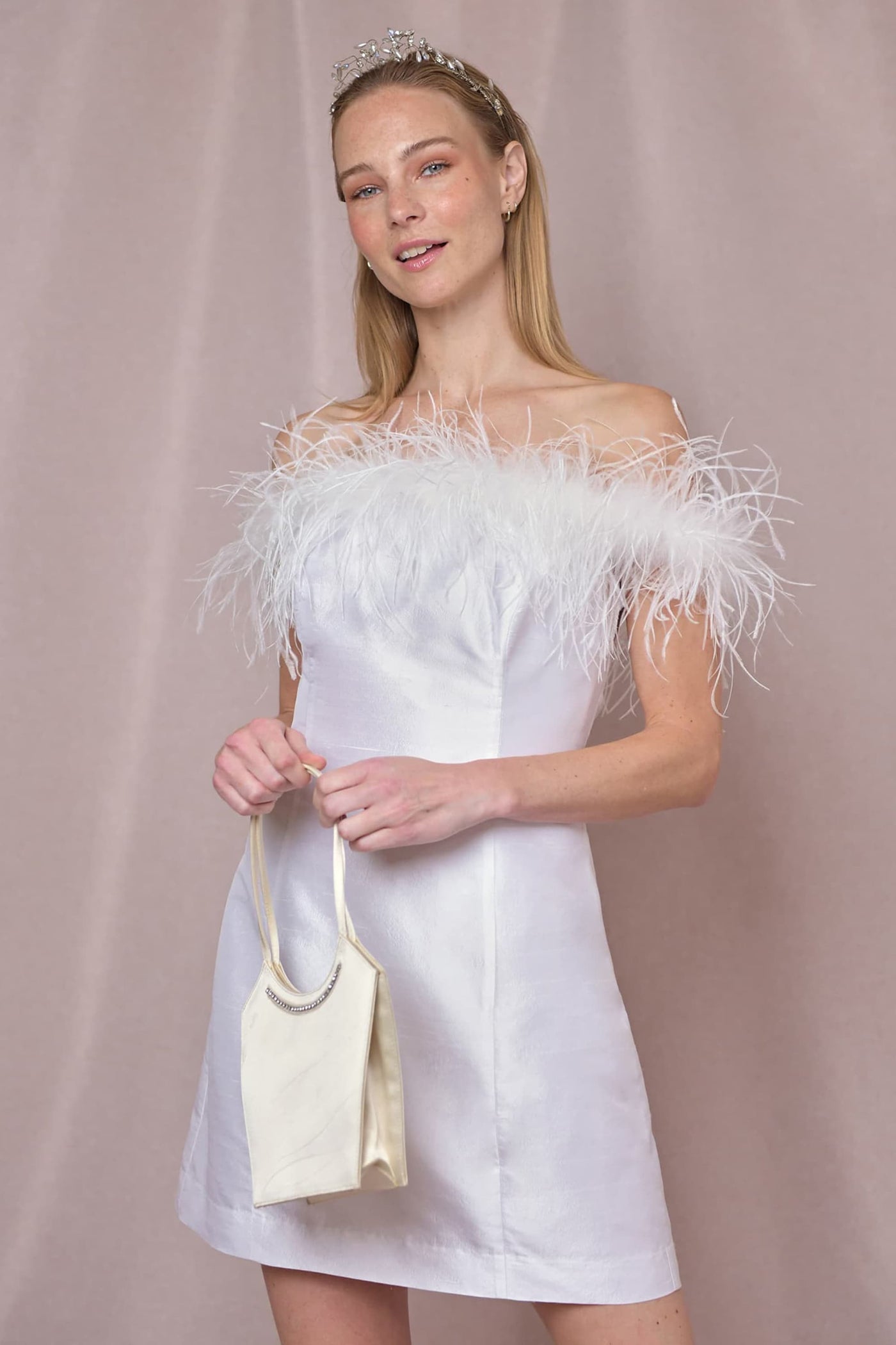 A woman smiles softly at the camera, wearing Lena - Feather Ivory, a white silk-jacquard mini dress with a Bardot neckline and feather trim. She pairs it with a small cream handbag and a silver tiara against a light beige background.