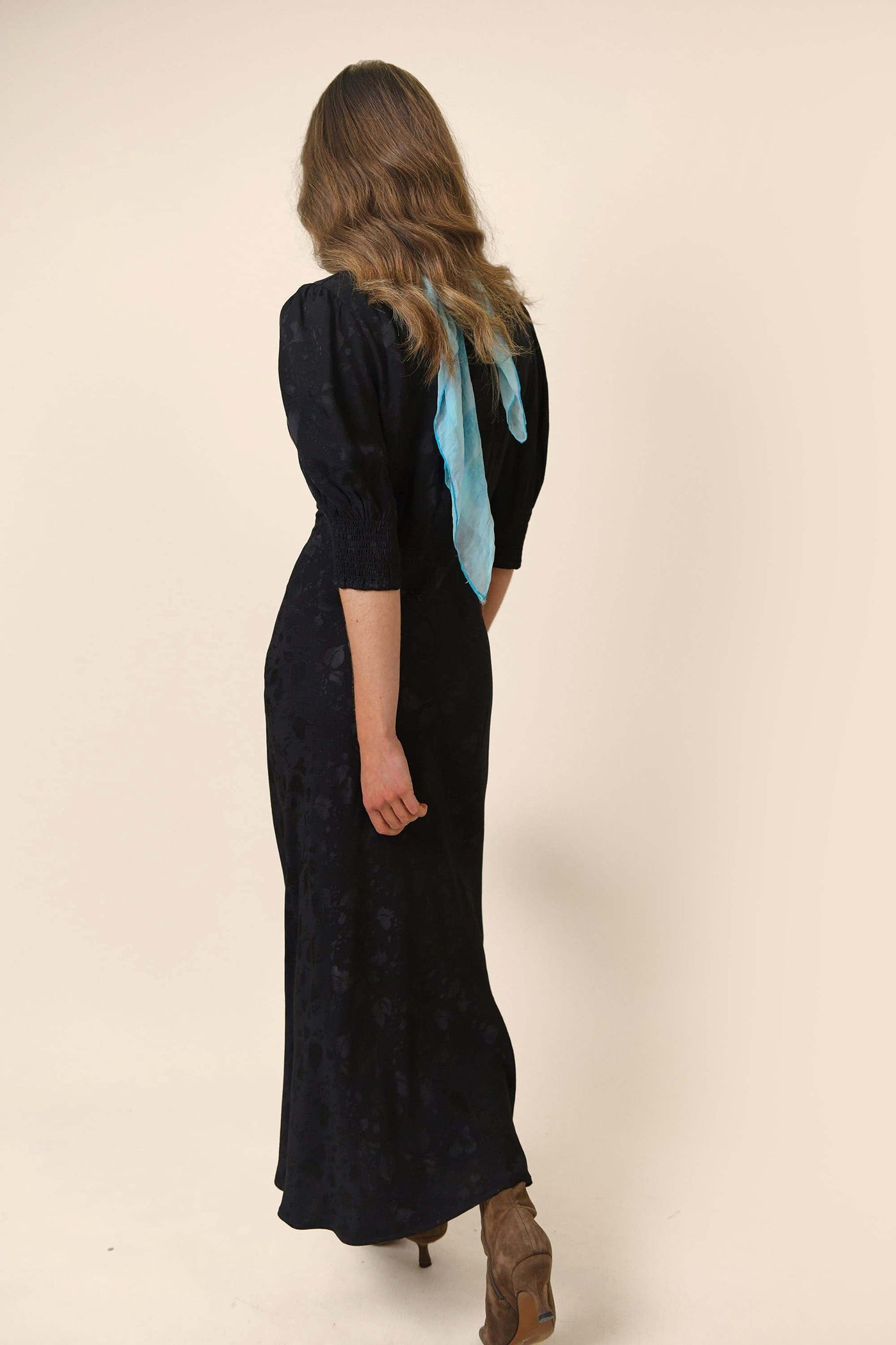 A woman with wavy light brown hair, wearing the RIXO Zadie - Leaf Heart Black midi dress and tan ankle boots, stands facing away from the camera with a blue scarf draped down her back against a plain light background.