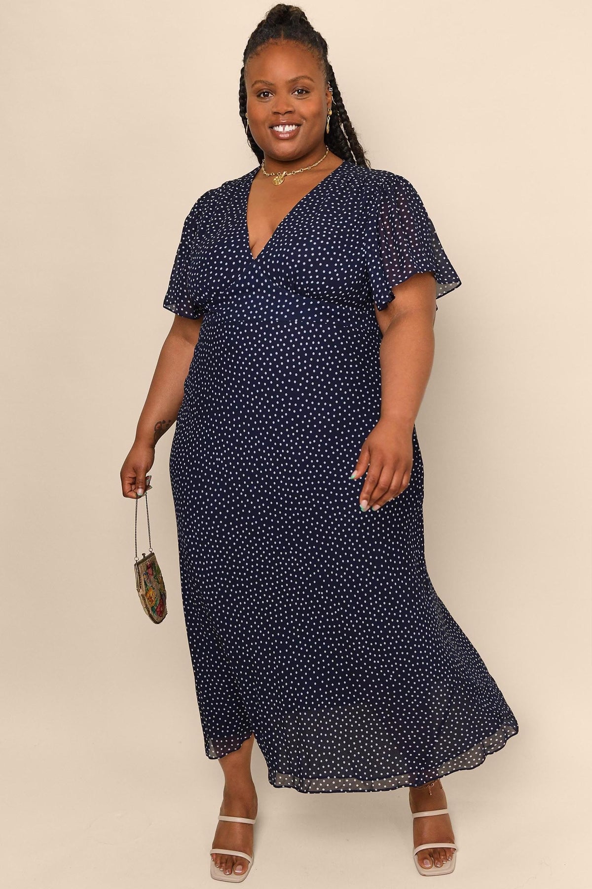 A smiling woman stands against a beige background wearing the Florida - Mini Dot Navy fit and flare maxi dress with short sleeves, white sandals, and holding a small purse.